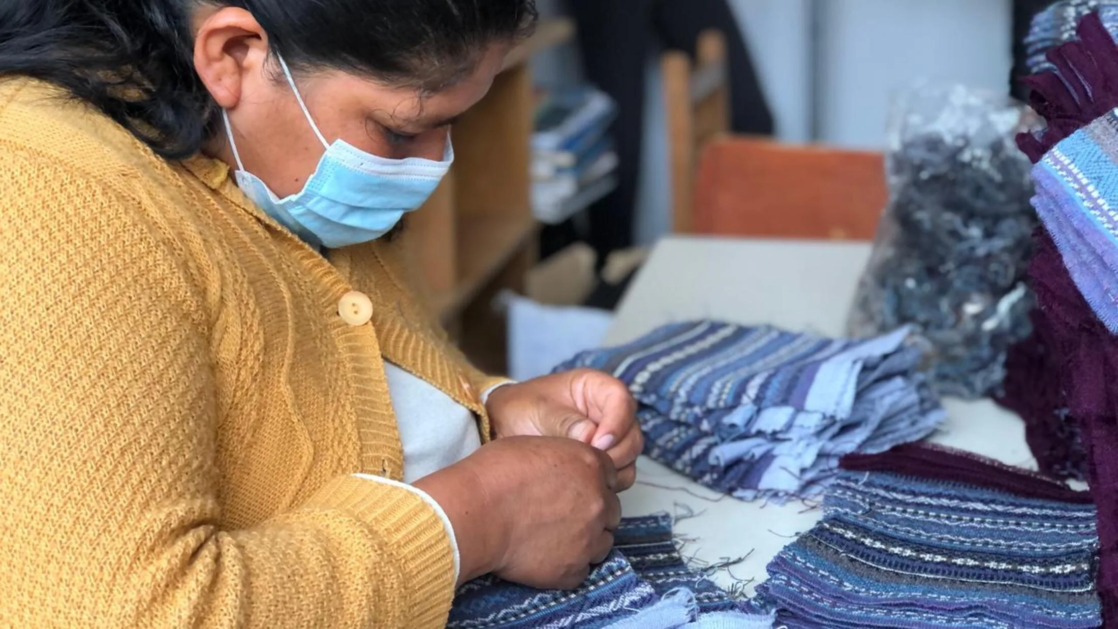 Una trabajadora del taller, con chompa mostaza, rematando a mano prendas a rayas.